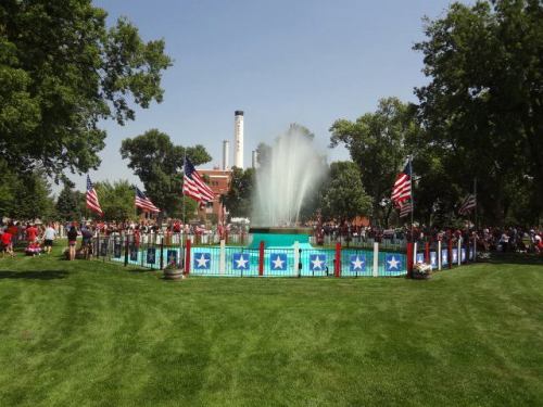 A festive gathering around a fountain, surrounded by American flags and greenery, on a sunny day.