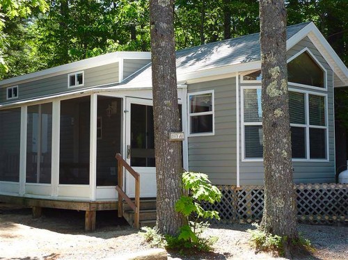 A small gray house with a screened porch, surrounded by trees and gravel, set in a wooded area.