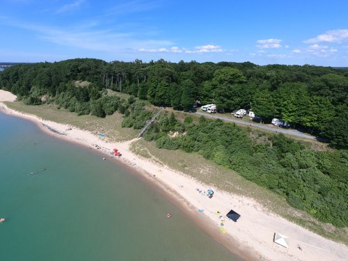 Aerial view of a beach with people, tents, and trees along the shore, surrounded by clear blue water and a green landscape.