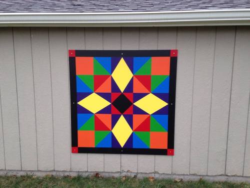A colorful geometric quilt pattern displayed on a gray wall, featuring stars and triangles in vibrant colors.