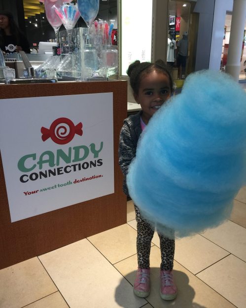 A child holds a large blue cotton candy in front of a "Candy Connections" sign in a shopping area.