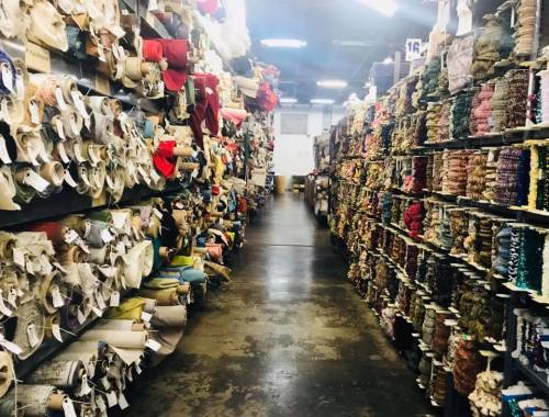 A wide aisle between shelves filled with colorful rolls of fabric and spools of thread in a textile warehouse.