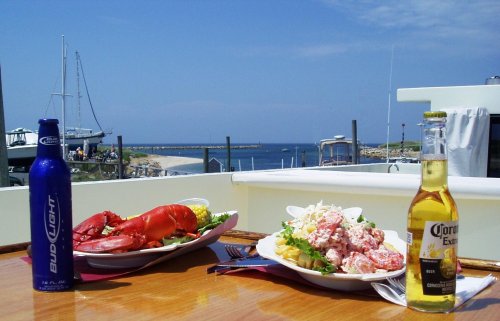 A scenic view of a seafood meal with lobster and drinks, overlooking a marina and blue sky.