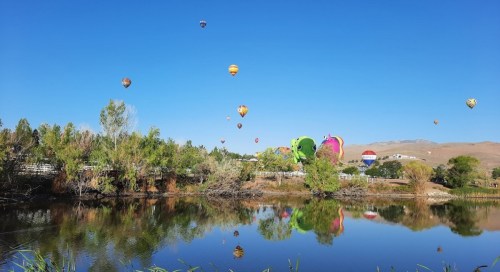 Colorful hot air balloons float over a serene lake, surrounded by lush greenery and clear blue skies.
