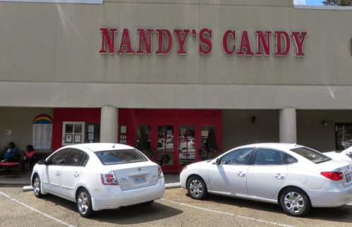 Exterior of Nandy's Candy shop with a red sign, featuring two parked white cars in front.