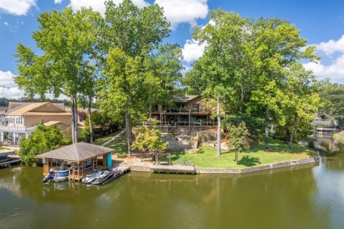 A scenic view of a lakeside home surrounded by trees, with a dock and jet skis on the water. Bright blue sky above.