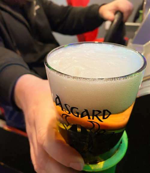 A close-up of a frothy beer in a glass with "Asgard" printed on it, held by a person in a dark shirt.