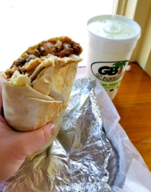 A hand holding a large burrito next to a cup of drink on a wooden table.