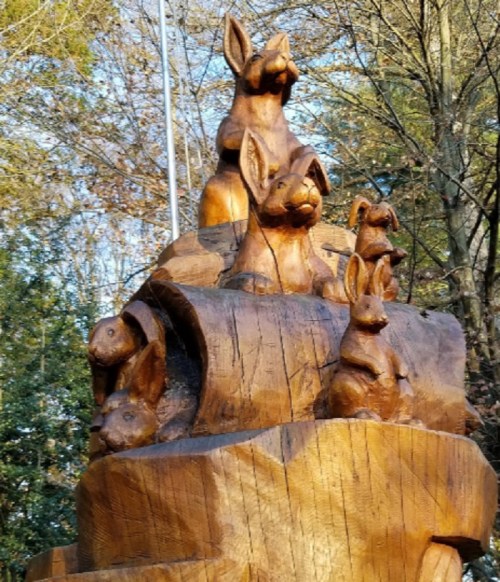 Wooden sculpture of several rabbits stacked on a log, surrounded by trees and natural scenery.