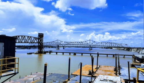 A view of a river with a bridge in the background, under a bright blue sky with scattered clouds.