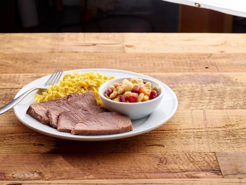 A plate with sliced beef, egg noodles, and a small bowl of mixed vegetables on a wooden table.
