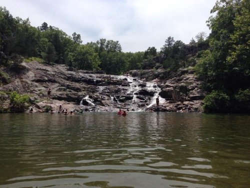 A serene waterfall cascades over rocks into a calm pool, surrounded by lush greenery and people enjoying the water.