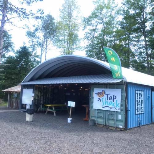 A blue metal building with a curved roof, featuring a sign for "Tap Shack" and surrounded by trees.