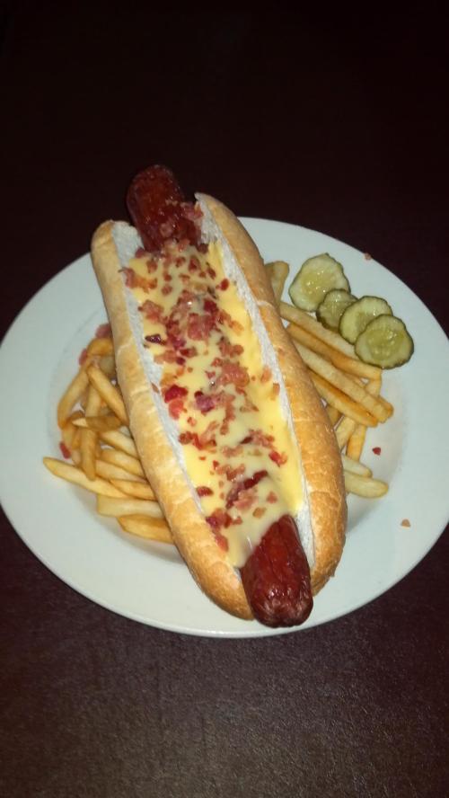 A hot dog in a bun topped with melted cheese and bacon bits, served with fries and pickles on the side.