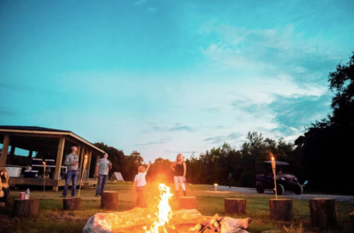 A group of people gathered around a campfire at dusk, with a wooden structure and trees in the background.