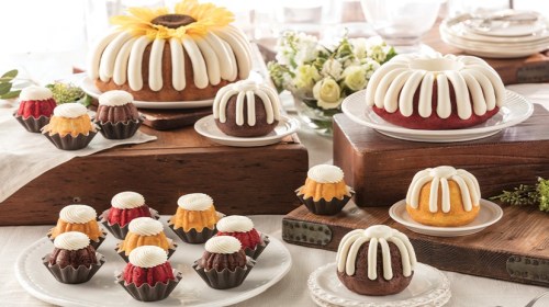 A variety of colorful bundt cakes and mini bundtlets with cream frosting, displayed on elegant plates and wooden stands.