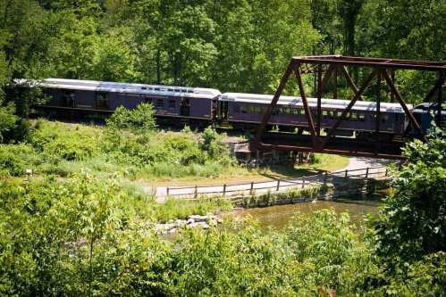 A vintage train crosses a red metal bridge over a river, surrounded by lush green trees and foliage.