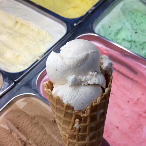 A hand holding a cone with two scoops of white ice cream, surrounded by various colorful ice cream flavors in tubs.