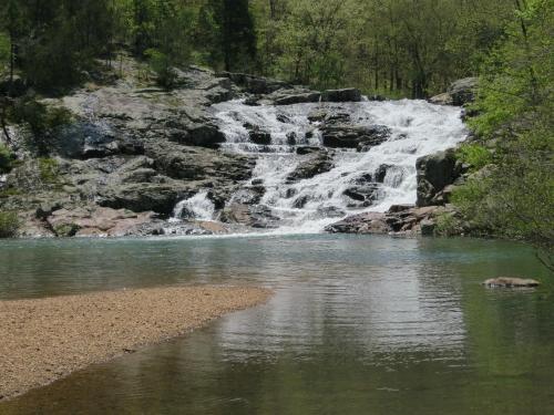 A serene waterfall cascades over rocks into a calm pool, surrounded by lush greenery and a sandy shore.