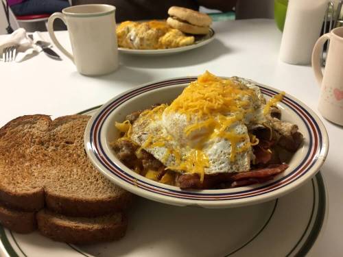 A hearty breakfast bowl with eggs, cheese, bacon, and toast on the side, served with coffee in the background.