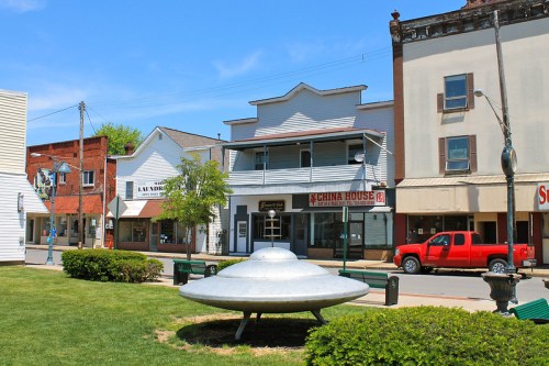 A metallic UFO sculpture sits on a grassy area in a small town, with shops and a red truck in the background.