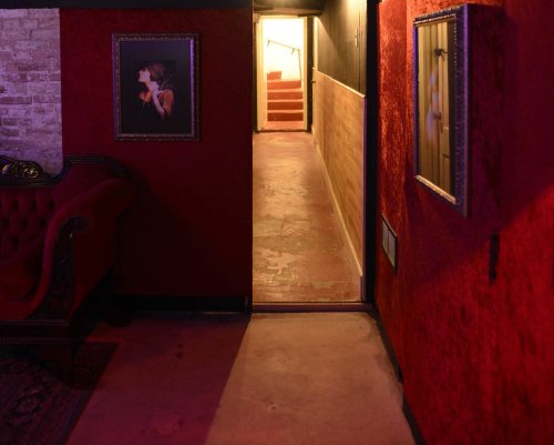 A dimly lit hallway with red walls, featuring framed portraits and a staircase at the end. A vintage couch is visible.