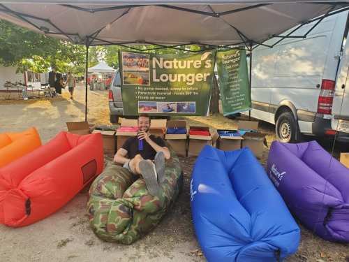 A man relaxes on a colorful inflatable lounger at a market stall, surrounded by various loungers and a display banner.
