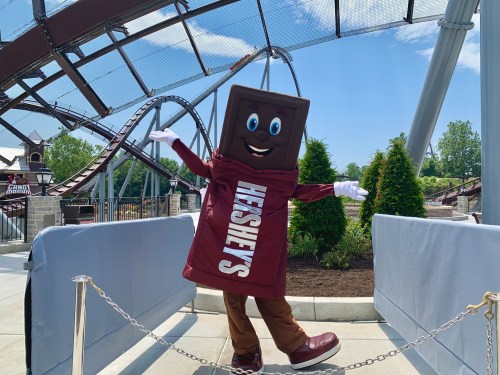 A cheerful chocolate bar mascot poses with arms outstretched in front of a roller coaster at an amusement park.