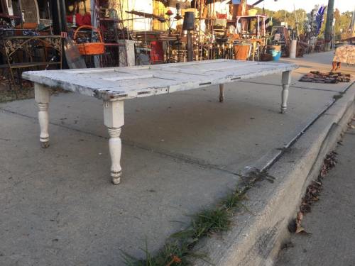A weathered white table with four legs, set on a sidewalk surrounded by various vintage items and furniture.