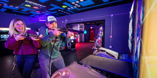 Two people joyfully playing a shooting arcade game, with colorful lights and retro decor in the background.
