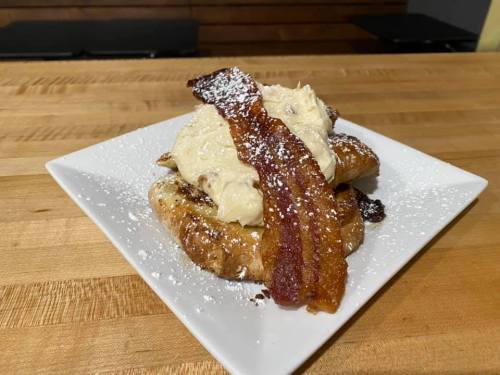 A plate of French toast topped with creamy frosting and crispy bacon, dusted with powdered sugar.