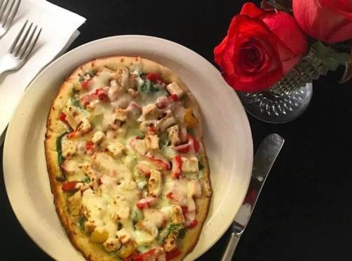 A delicious pizza topped with chicken, peppers, and melted cheese, next to a vase of red roses on a black table.