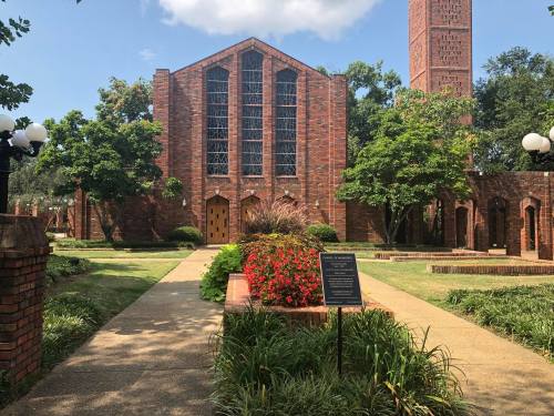 A brick church with large windows, surrounded by greenery and flowers, under a blue sky with scattered clouds.