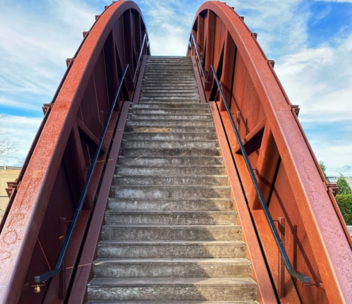 A steep, red metal bridge with a staircase leading up, set against a blue sky with scattered clouds.
