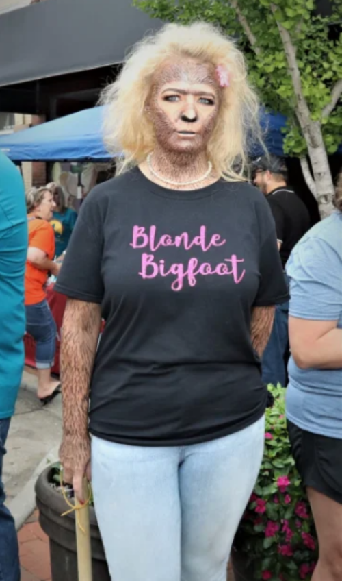 A person with a unique appearance wearing a "Blonde Bigfoot" t-shirt, standing outdoors among a crowd.