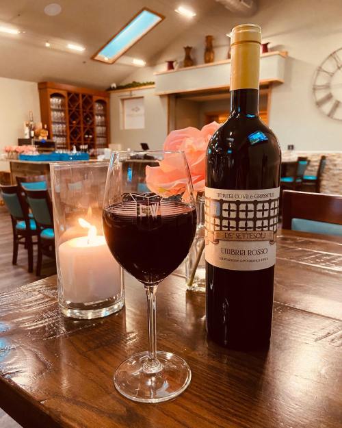 A glass of red wine beside a candle and a bottle of Umbra Rosso on a wooden table in a cozy setting.
