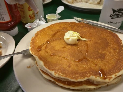 A stack of golden pancakes topped with butter and syrup, served on a white plate with condiments in the background.