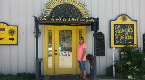 A woman stands outside a bright yellow restaurant entrance with a sunflower design, showcasing a welcoming sign.
