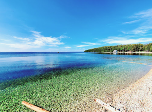 A serene lake view with clear water, a sandy shore, and lush green trees under a bright blue sky.