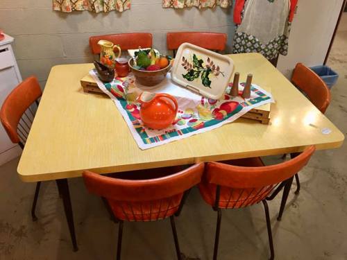 A retro dining table with orange chairs, adorned with a colorful tablecloth and a bowl of fruit.