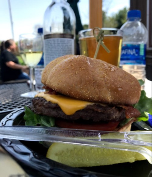 A close-up of a cheeseburger with lettuce, tomato, and a pickle, set on a table with drinks in the background.