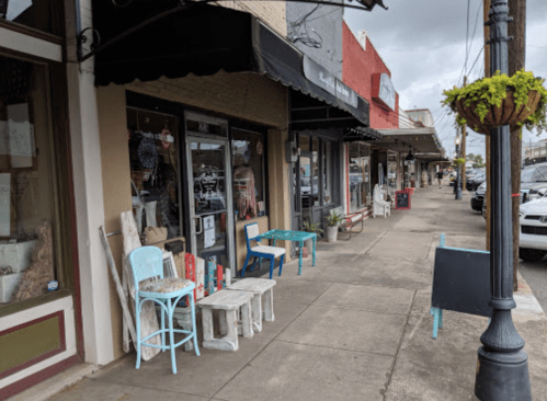 A quaint street scene featuring shops with outdoor seating and colorful chairs along a sidewalk.