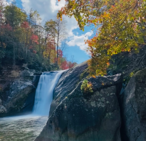 A serene waterfall cascades over rocks, surrounded by colorful autumn foliage and a bright blue sky.