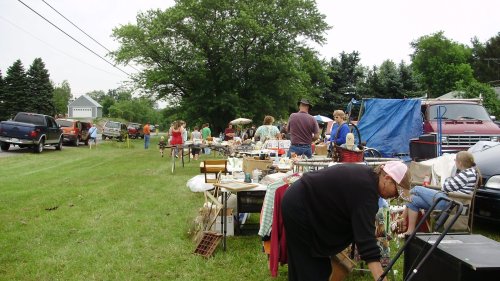 A bustling outdoor flea market with vendors and shoppers browsing tables filled with various items. Green grass and trees in the background.