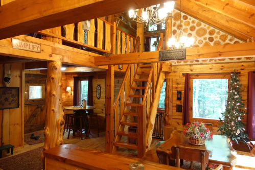 Cozy wooden cabin interior with a staircase, dining area, and a Christmas tree, featuring warm lighting and rustic decor.