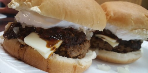 Two burgers on soft buns, featuring a beef patty, melted cheese, and a rich sauce, topped with onions and a dollop of cream.
