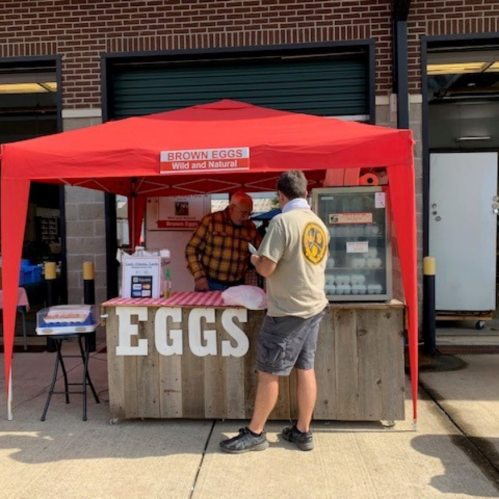 A vendor under a red tent sells brown eggs at a market, with a customer inquiring at the booth.
