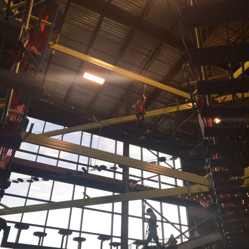 A person navigates a high ropes course, suspended on beams inside a large indoor facility with windows.