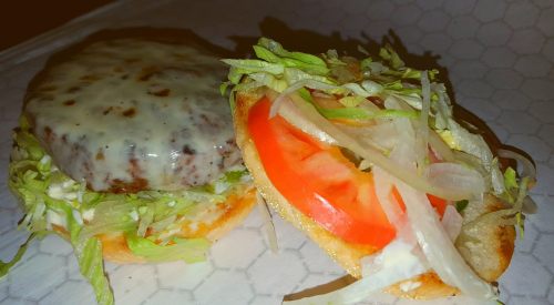 A close-up of a burger bun with melted cheese, lettuce, tomato, and onions on one half, and the other half empty.