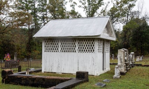 A small white structure with a metal roof in a cemetery, surrounded by gravestones and trees.